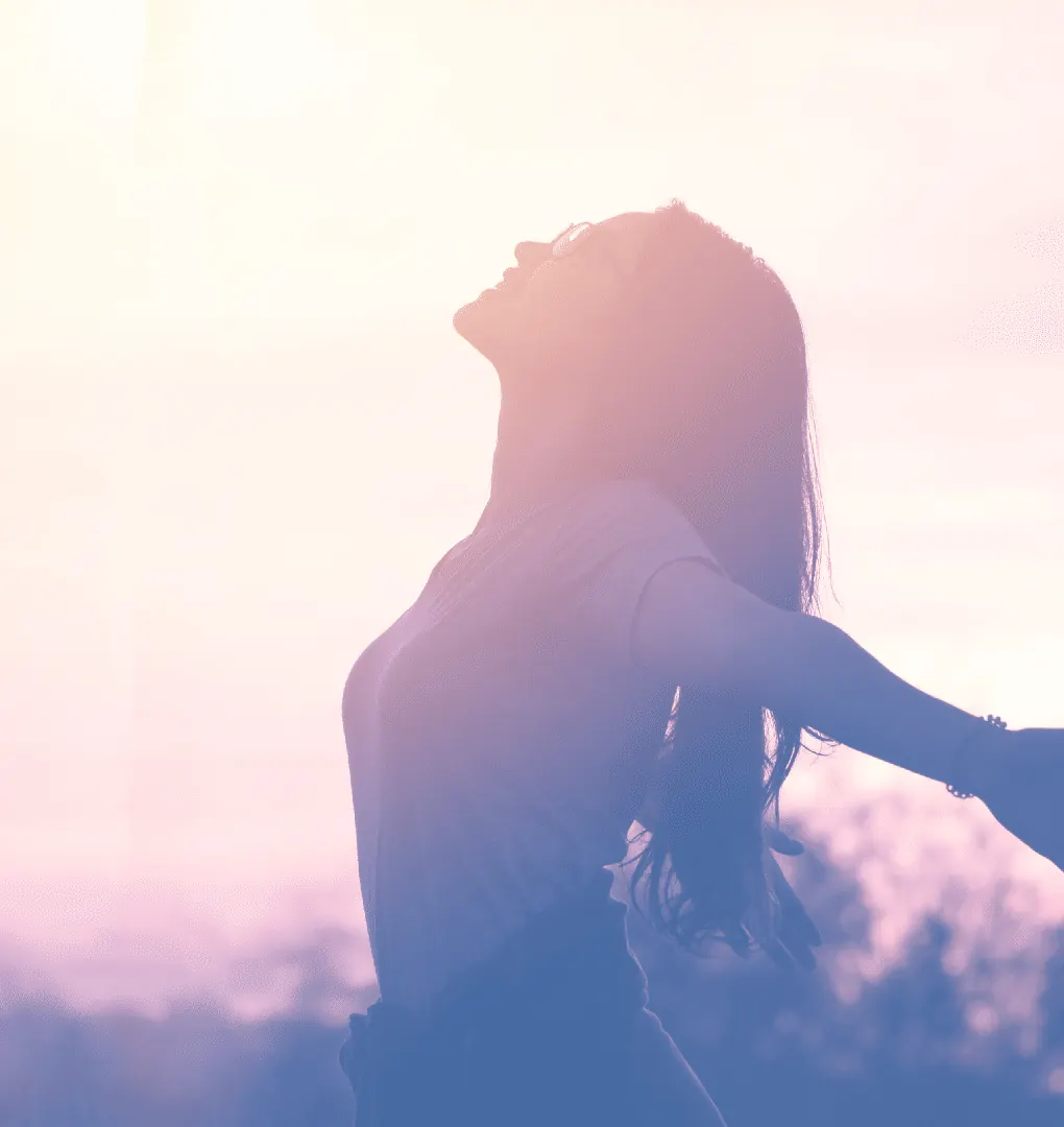 Una mujer, de perfil y con los brazos abiertos, mira al cielo en un amanecer de tonos pastel.
