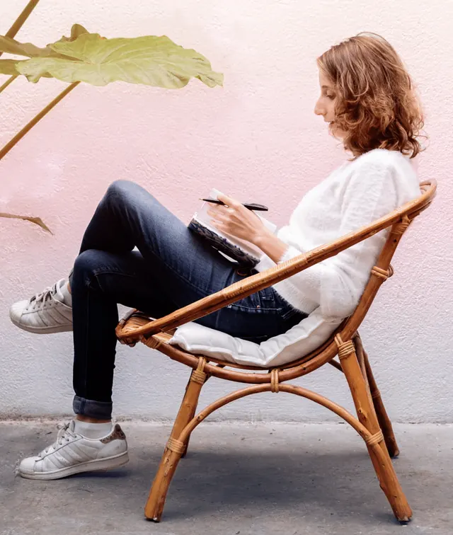 María del Mar Cinto, sentada en una butaca de mimbre, escribe en una libreta junto a una pared rosa.
